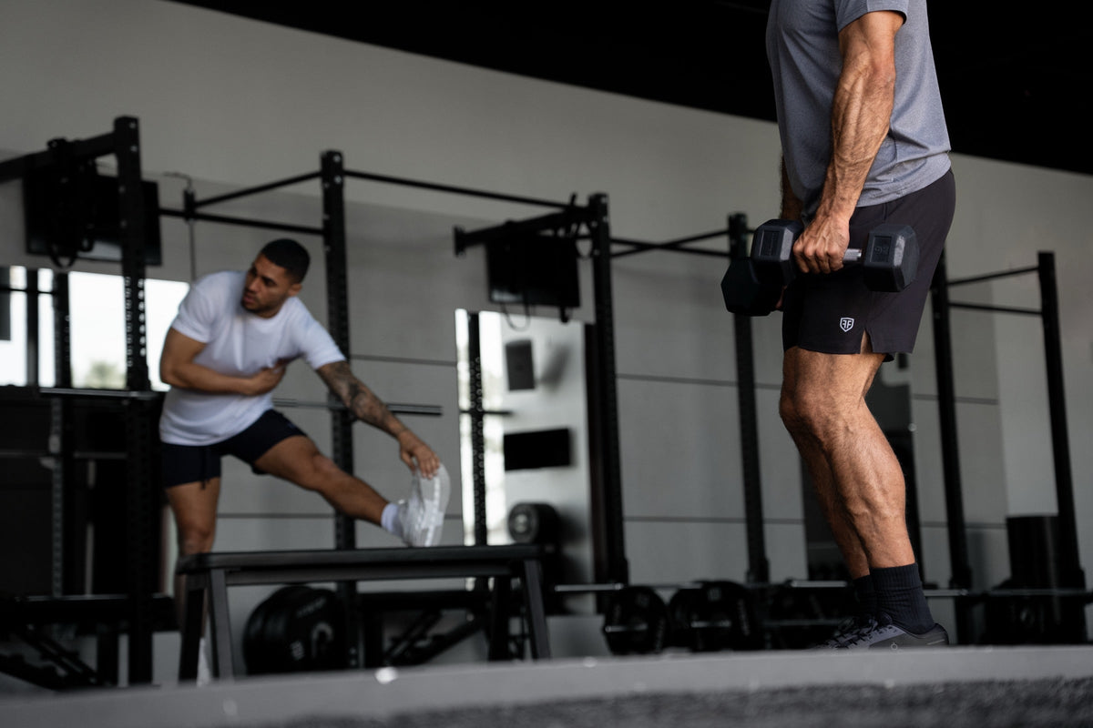 Two dads training at the gym in Father Fortified gear, one stretching and one carrying dumbbells